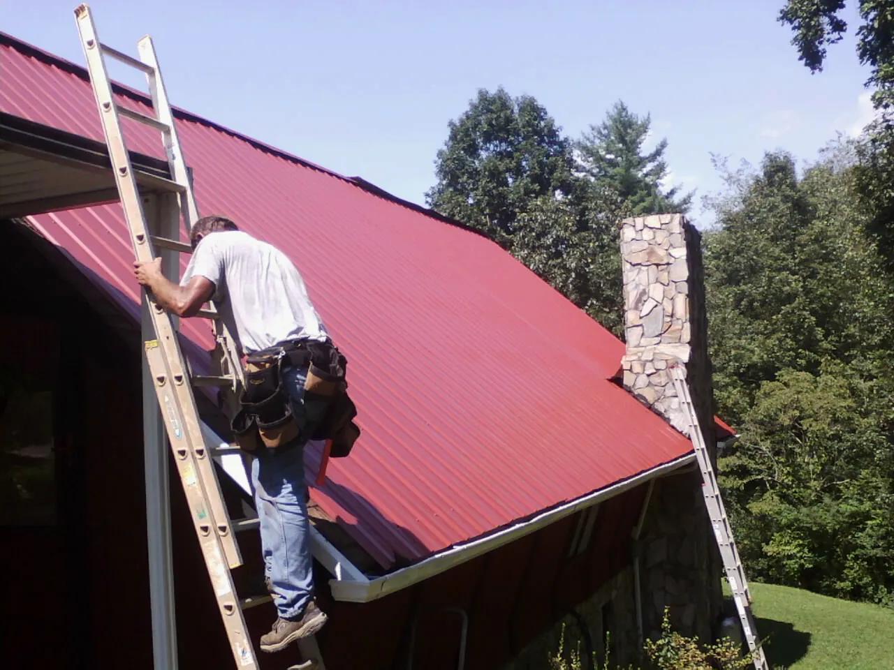Roofer on a ladder beside a red metal roof and stone chimney — Soffit & Fascia Repair by Roofing Brothers in Lutts